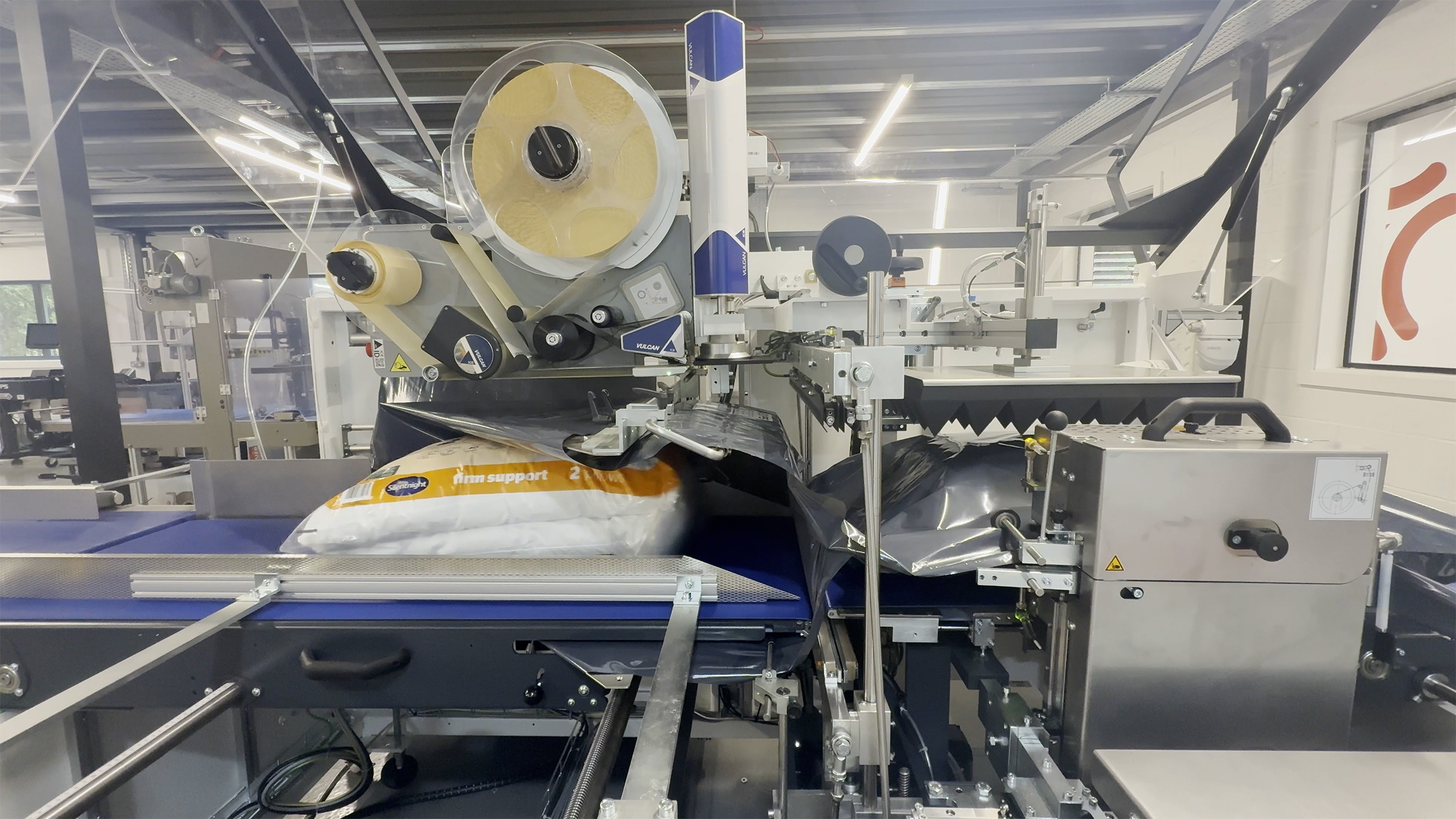 In an industrial manufacturing setting, a large roll of bright yellow material is being unwound and fed into a blue automated production machine with rollers, guides, and control panels; the material appears to be a thin film or adhesive tape moving through the processing line under overhead lighting.